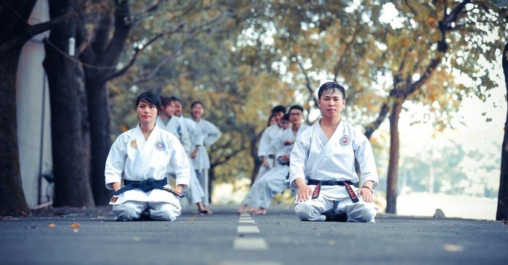 A man and a woman in white karate uniforms with black belts kneel on an outdoor path while other students stand behind them.