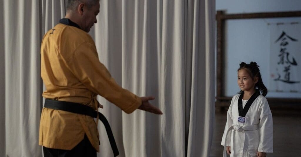 A karate instructor wearing a gold uniform and black belt reaches out his hand to a young girl in a white uniform during a class.