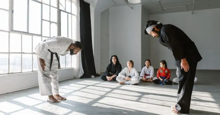 Two karate instructors bow to each other in a bright studio while a small group of students sits cross-legged on the floor watching.
